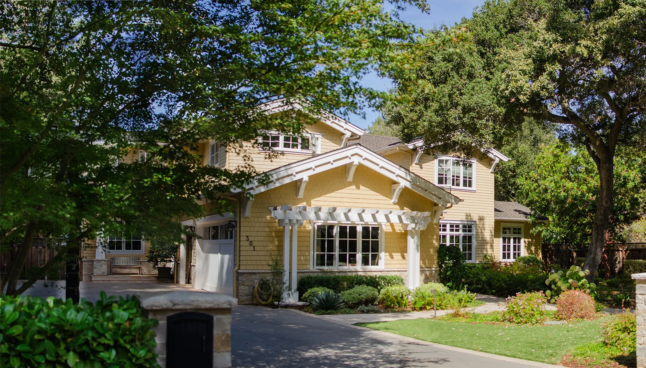 Driveway view of a Menlo Park residence with yellow siding, white trim, a pergola, and a stone base.