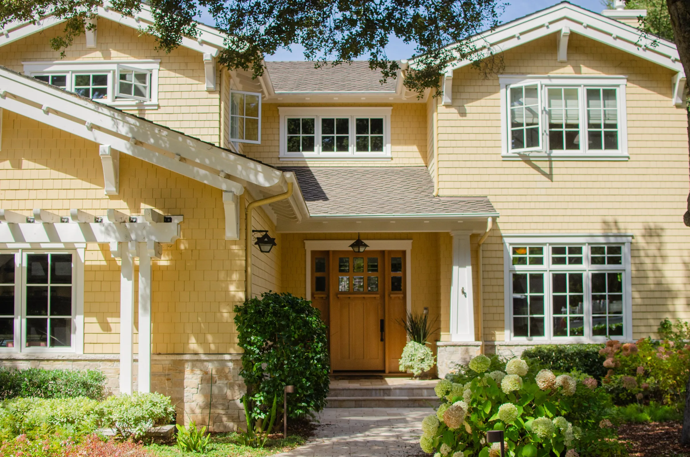 Front of a Menlo Park residence with yellow shingle siding, white trim, a wood door, and a white pergola over a stone base.