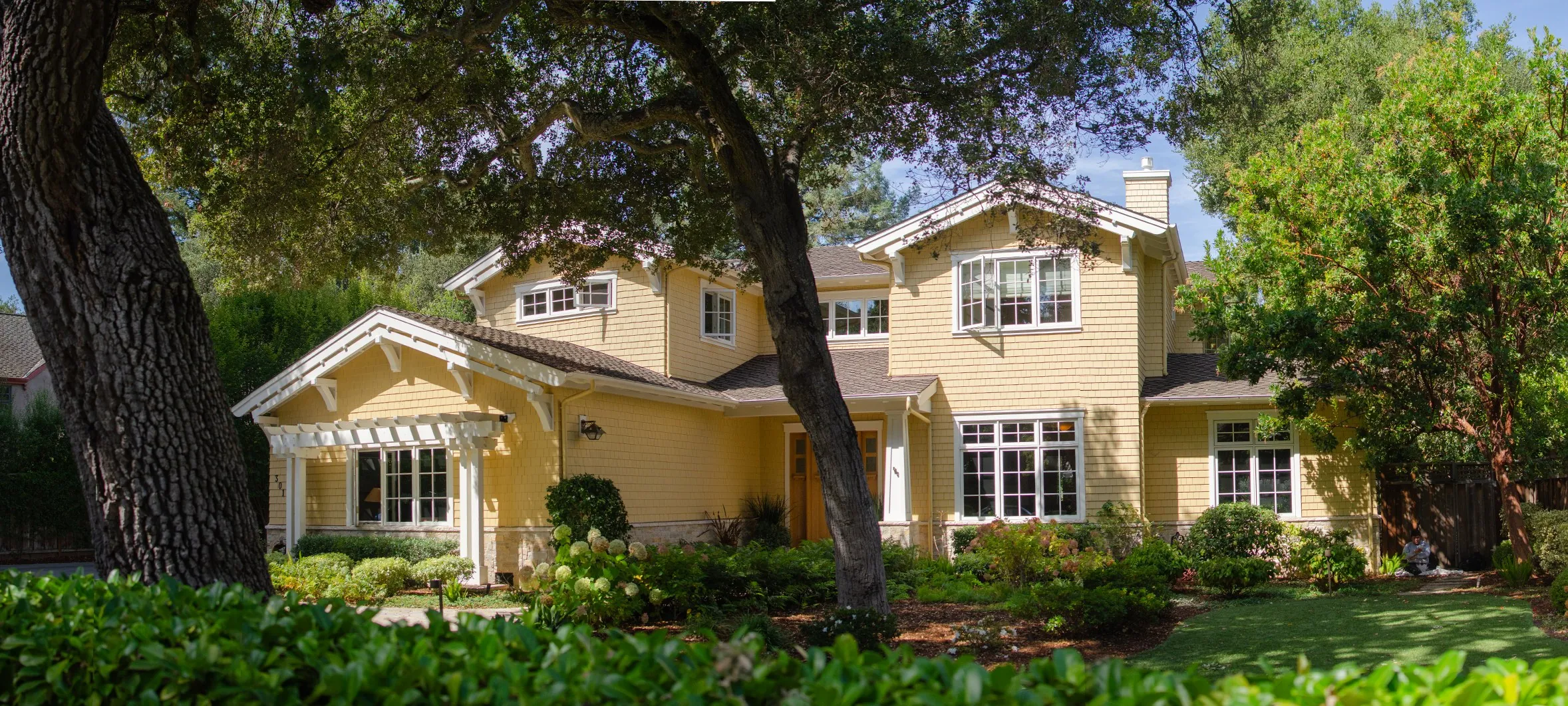 Street view of a Menlo Park residence with yellow shingle siding, white trim, a wood door, and a pergola, framed by trees.
