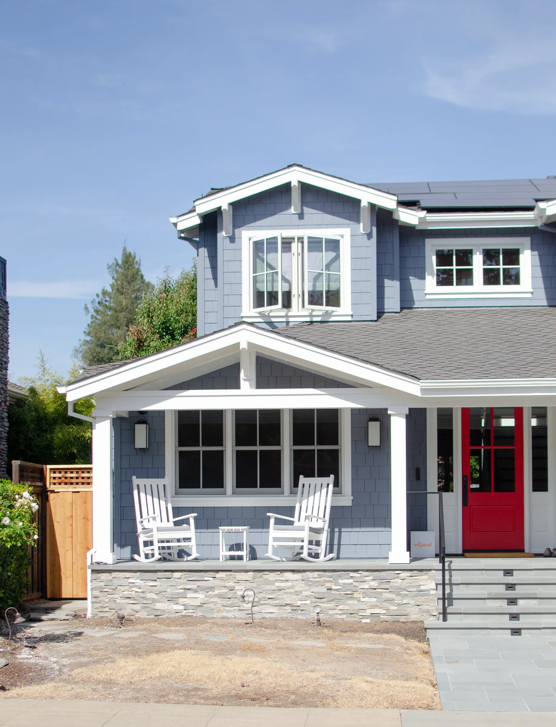 Covered front porch of a San Carlos residence with blue shingle siding, white columns and railings, a red entry door, and a stone porch base.